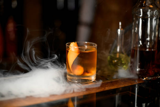 Golden cocktail in the glass with one ice cube and orange zest on the bar counter in the dark blurred background in the smoke